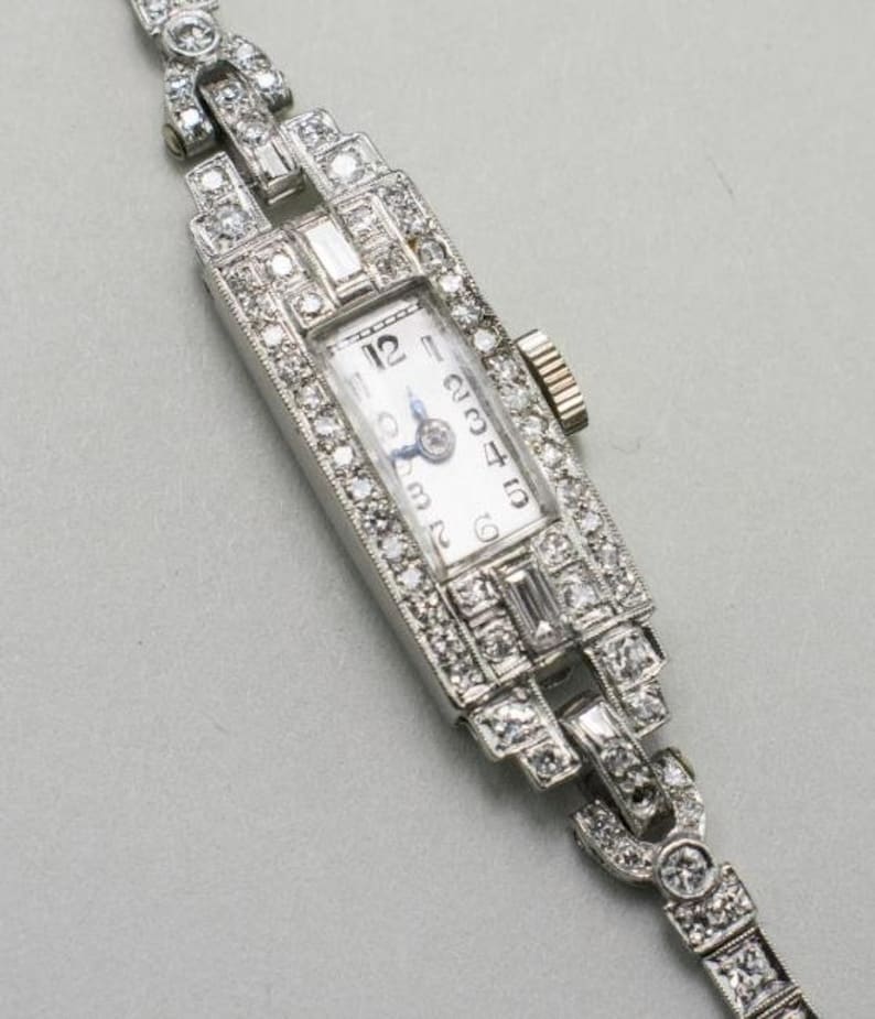 May include: A vintage rectangular wristwatch with a white face and silver hands. The watch is encrusted with numerous small diamonds, and the band is also adorned with diamonds. The watch has a classic, elegant design.