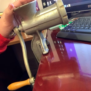 May include: A vintage, silver-colored meat grinder with a wooden handle. The grinder is clamped to a red surface, showcasing its design and functionality. The handle is made of wood and is a light brown color.