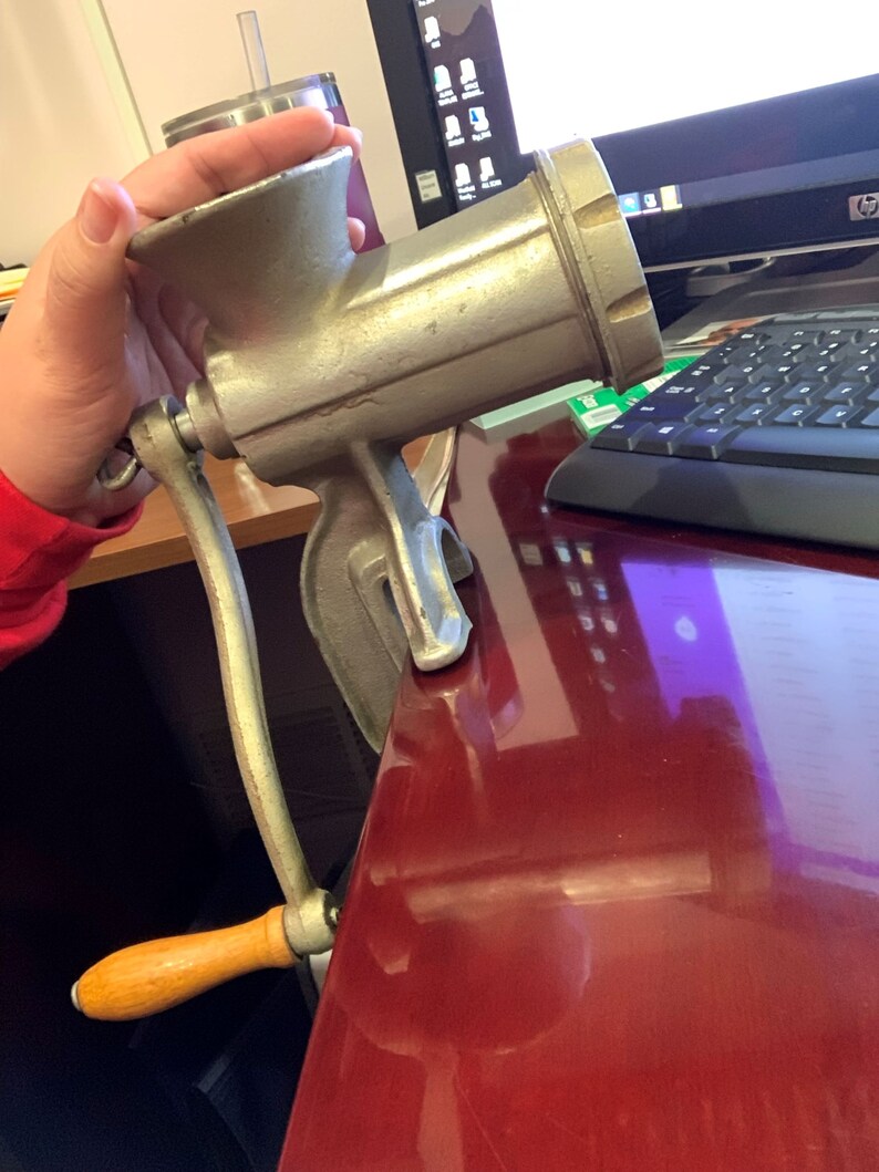 May include: A vintage, silver-colored meat grinder with a wooden handle. The grinder is clamped to a red surface, showcasing its design and functionality. The handle is made of wood and is a light brown color.