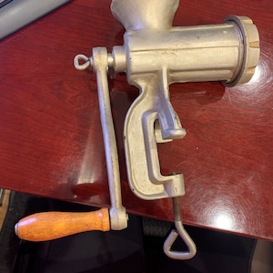 May include: A vintage, silver-colored meat grinder with a wooden handle. The grinder has a clamp to secure it to a surface and a crank for operation. The design is classic and functional, suggesting a tool for home food preparation.