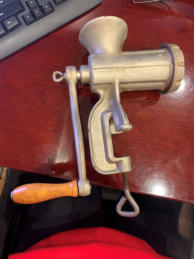 May include: A vintage, silver-colored meat grinder with a wooden handle. The grinder has a clamp to secure it to a surface and a crank for operation. The design is classic and functional, suggesting a tool for home food preparation.