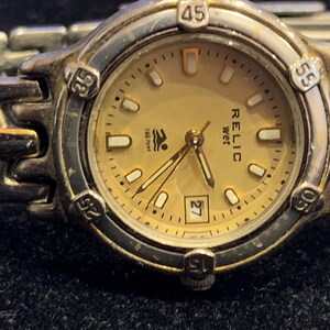 Peut inclure: Une montre-bracelet en acier inoxydable couleur or avec un cadran or et des aiguilles noires. Le cadran de la montre comporte une fen&ecirc;tre de date et le texte "RELIC wet 165 feet".