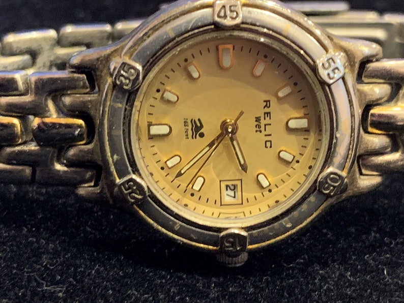 Peut inclure: Une montre-bracelet en acier inoxydable couleur or avec un cadran or et des aiguilles noires. Le cadran de la montre comporte une fen&ecirc;tre de date et le texte "RELIC wet 165 feet".