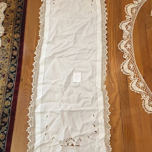 May include: White crocheted table runner with delicate floral embroidery and a lace border. The runner is approximately 50 inches long and 14 inches wide.