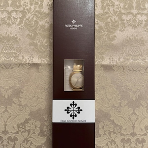 May include: A brown box with the Patek Philippe Geneva logo and a gold watch inside. The box also has a white label with the Patek Philippe logo and the text "HSWA Customer Service".