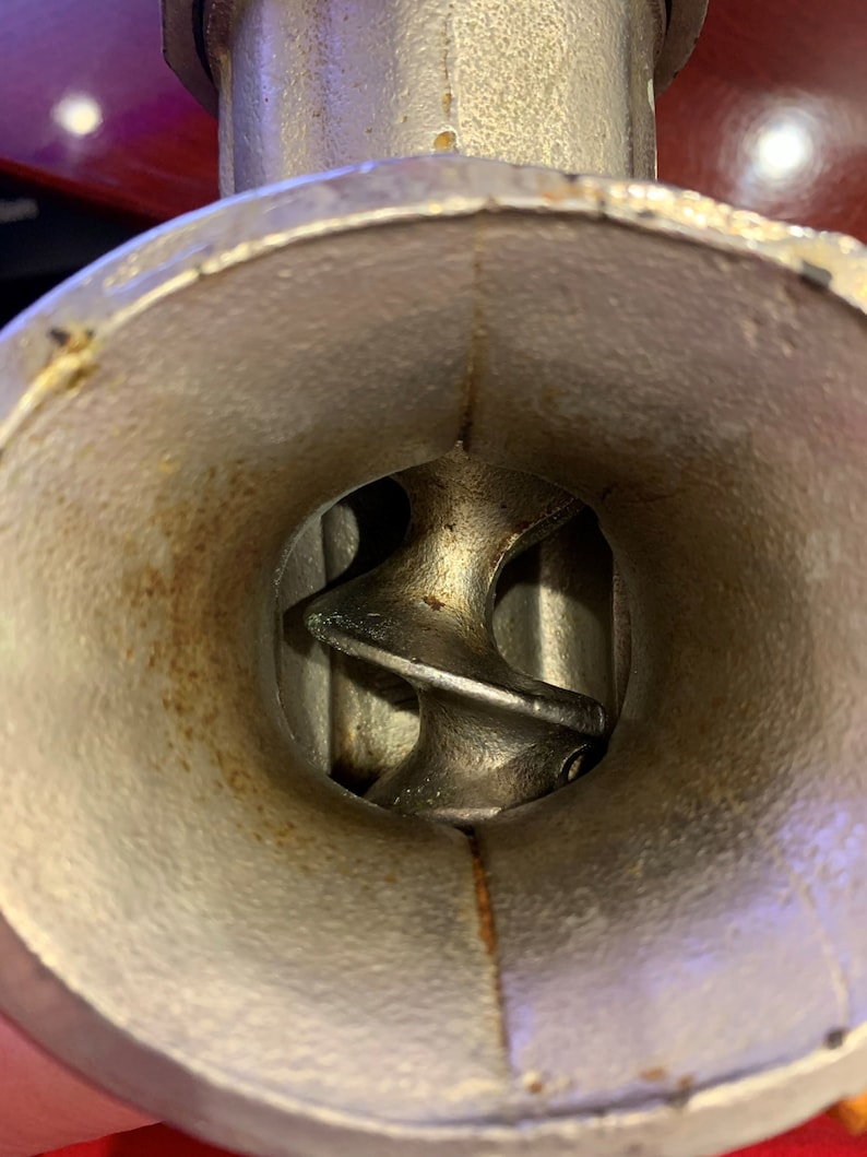 May include: Close-up of a vintage metal meat grinder. The interior shows the auger and the inside of the grinder. The metal has a worn, aged appearance with some rust. The grinder is designed for food preparation.