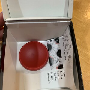 Puede incluir: Una pieza de rompecabezas redonda roja se encuentra en una caja blanca con las palabras "Mitte, Milieu, Centro, Midden, Middle, En medio" impresas en una tarjeta blanca.