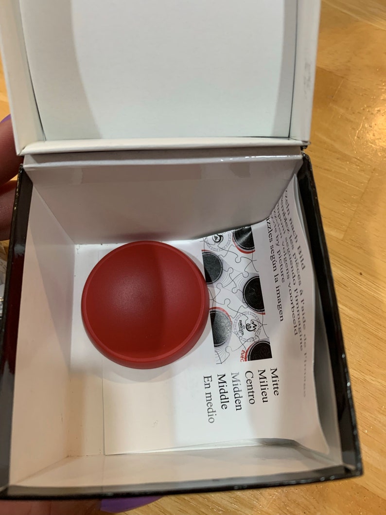 Puede incluir: Una pieza de rompecabezas redonda roja se encuentra en una caja blanca con las palabras "Mitte, Milieu, Centro, Midden, Middle, En medio" impresas en una tarjeta blanca.