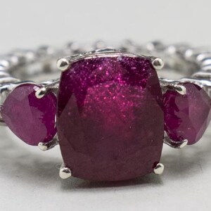 May include: A silver ring with three large, deep red gemstones. The center stone is a square cushion cut, and the two side stones are heart-shaped.