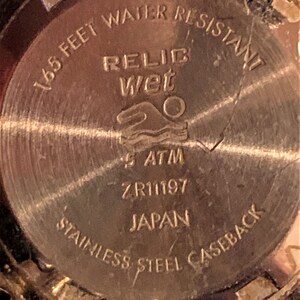 Peut inclure: Le fond d'un bo&icirc;tier de montre dor&eacute; avec le texte "165 feet water resistant", "Relic Wet", "5 ATM", "ZR11197", "Japan", et "Stainless Steel Caseback".