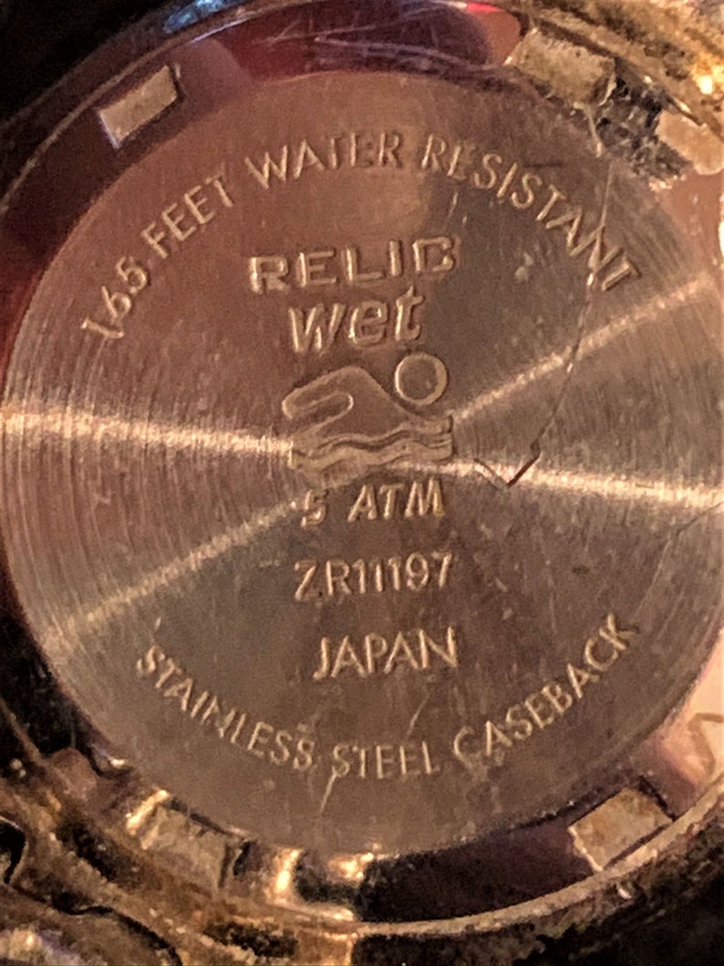Peut inclure: Le fond d'un bo&icirc;tier de montre dor&eacute; avec le texte "165 feet water resistant", "Relic Wet", "5 ATM", "ZR11197", "Japan", et "Stainless Steel Caseback".
