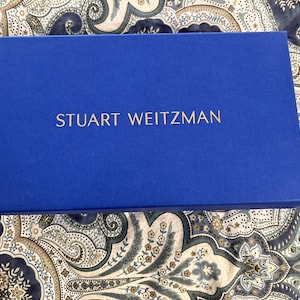 Könnte beinhalten: Eine blaue Box mit dem Text "STUART WEITZMAN" in goldenen Buchstaben.