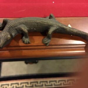 May include: A bronze lizard figurine with a textured, scaly surface. The lizard is posed with its head turned to the side and its tail curled up.