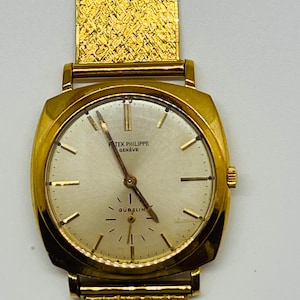 May include: A gold Patek Philippe Geneve wristwatch with a square face and a gold band. The watch face has a white background with black Roman numeral hour markers and black hands. The watch face also has the text "Patek Philippe Geneve" and "Gubelin" printed on it.