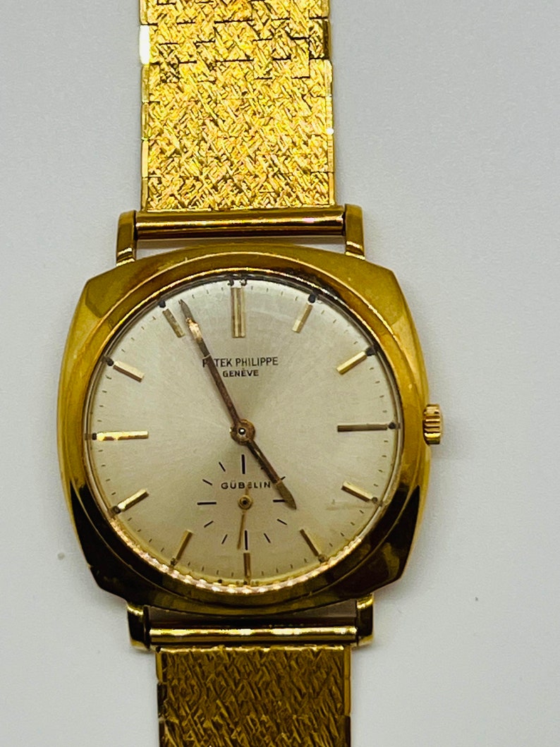 May include: A gold Patek Philippe Geneve wristwatch with a square face and a gold band. The watch face has a white background with black Roman numeral hour markers and black hands. The watch face also has the text "Patek Philippe Geneve" and "Gubelin" printed on it.