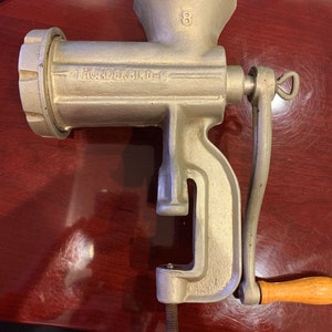 May include: A vintage silver-colored Thunderbird meat grinder with a wooden handle. The grinder has the number "8" on the top and the brand name is printed on the side. It is designed to clamp onto a table.
