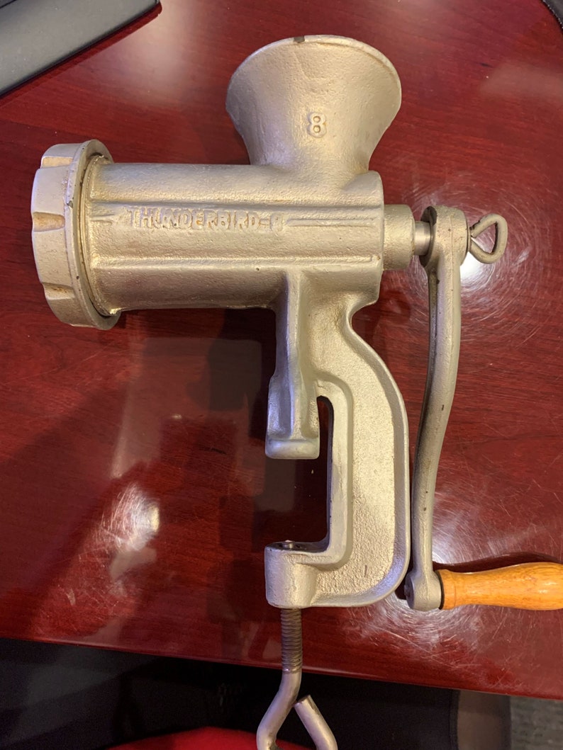 May include: A vintage silver-colored Thunderbird meat grinder with a wooden handle. The grinder has the number "8" on the top and the brand name is printed on the side. It is designed to clamp onto a table.
