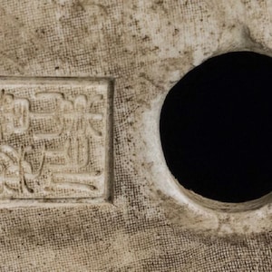 Puede incluir: Primer plano de una superficie texturizada blanquecina con un agujero circular y un dise&ntilde;o cuadrado en relieve con caracteres asi&aacute;ticos. La superficie tiene una apariencia tejida, con un fondo oscuro.