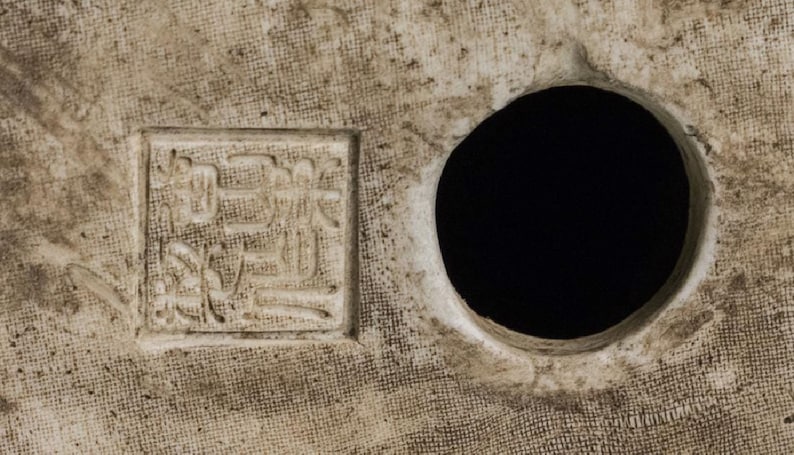 Puede incluir: Primer plano de una superficie texturizada blanquecina con un agujero circular y un dise&ntilde;o cuadrado en relieve con caracteres asi&aacute;ticos. La superficie tiene una apariencia tejida, con un fondo oscuro.