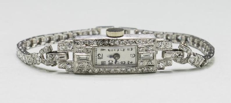 May include: A vintage rectangular wristwatch with a silver band and face. The watch face is white with black numerals and hands. The watch is encrusted with small, sparkling gemstones. The band is made of small, connected links.