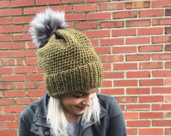 May include: A green crocheted beanie with a fluffy gray pom pom. The beanie is worn by a person with blonde hair and a black jacket.