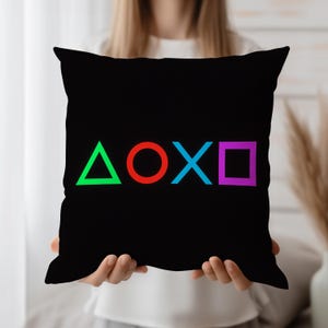 May include: Black square pillow featuring the green triangle, red circle, blue X, and purple square symbols. The symbols are arranged horizontally across the center of the pillow. The pillow is held up by a person.
