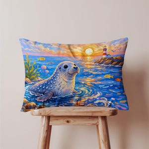 May include: A rectangular pillow featuring a whimsical illustration of a seal in the ocean. The seal is light blue with dark spots, surrounded by blue water, yellow fish, and jellyfish. A lighthouse and sunset are in the background. The pillow is on a wooden stool.