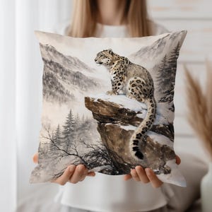May include: A decorative pillow featuring a snow leopard perched on a snowy rock, with a mountainous winter landscape in the background. The pillow has a soft, neutral color palette with shades of gray, white, and brown.
