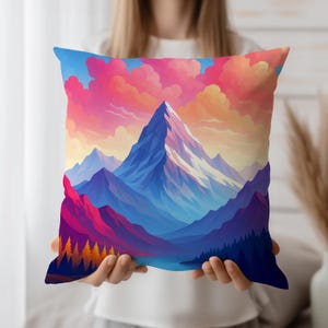 May include: Square cushion with a colourful mountain scene. The design features a snow-covered peak, blue and purple mountains, and a sky with pink and orange clouds. The cushion is held by a person.