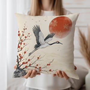 Op de afbeelding: Decoratief kussen met een Japans geïnspireerd ontwerp. Het heeft een vliegende kraanvogel, een rode zon en bloeiende takken. Het kleurenpalet omvat crème, rood, zwart en grijs.