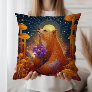 Puede incluir: Cojín decorativo cuadrado con un oso sosteniendo flores moradas, sobre un cielo estrellado con luna llena y setas estilizadas. El oso tiene pelaje naranja y está mirando hacia arriba. El cojín tiene un fondo azul oscuro.
