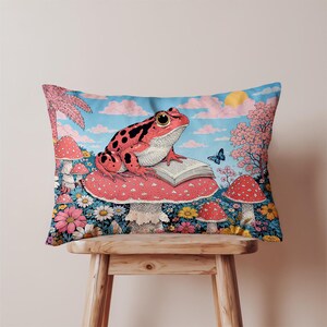 May include: A decorative pillow featuring a whimsical illustration of a red frog with black spots, sitting on a mushroom and reading a book. The pillow has a blue sky background with pink clouds, flowers, and other mushrooms.