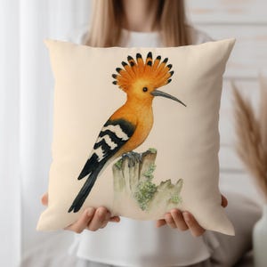 May include: A cushion with a watercolour illustration of a hoopoe bird perched on a tree stump. The bird has orange, black, and white markings with a distinctive crest. The cushion has a cream-coloured background.