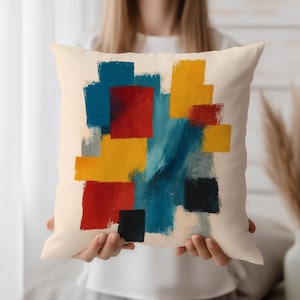 Puede incluir: Cojín cuadrado color crema con un diseño abstracto. El diseño presenta pinceladas superpuestas en rojo, amarillo, azul y negro. El cojín está sostenido por una persona.