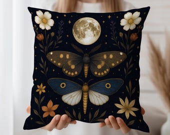 May include: Square decorative pillow with a dark blue background featuring a full moon, two moths, flowers, and stars. The moths are brown and blue with intricate details. Floral elements include white, orange, and yellow flowers.
