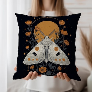 Könnte beinhalten: Quadratisches schwarzes Kissen mit einer detaillierten Illustration einer Motte mit orangefarbenen Punkten und einem großen orangefarbenen Mond im Hintergrund. Die Motte ist von orangefarbenen Blumen und dunkelgrünem Laub umgeben.