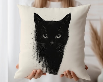 May include: Cream-colored square throw pillow featuring a black cat illustration. The cat's face is centered, with a stylized, artistic design. The cat's fur is black, and the eyes are a light green. The pillow is held up by a person.
