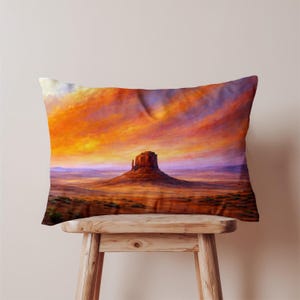 May include: Rectangular pillow featuring a vibrant landscape painting. The artwork depicts a mesa under a dramatic sunset with orange, yellow, and purple hues. The pillow is resting on a wooden stool.