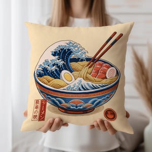 Op de afbeelding: Een vierkant beige kussen met een kleurrijke illustratie van een kom ramen met eetstokjes. De ramenkom is versierd met een blauw en oranje golfpatroon. De afbeelding bevat Japanse tekst.