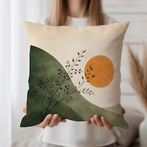 May include: Square decorative pillow with an abstract landscape design. The pillow features a green and beige color scheme with a large orange sun and black plant silhouettes. The pillow is held by a person.