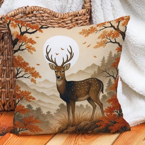 May include: Square decorative pillow featuring a deer in a forest scene. The artwork depicts a deer with large antlers standing in front of a full moon, surrounded by trees with orange leaves. The background shows mountains and a forest.