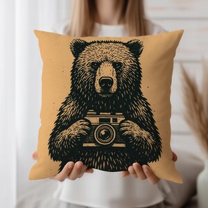 May include: Square decorative pillow with a black illustration of a bear holding a vintage camera. The bear is centered on a tan background. The pillow is a square shape and appears to be made of soft fabric.