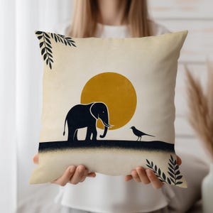 Könnte beinhalten: Dekoratives quadratisches Kissen mit minimalistischem Design. Es zeigt eine schwarze Elefanten-Silhouette, einen Vogel und eine gelbe Sonne vor beigem Hintergrund. Schwarze Blätter akzentuieren die Ecken. Das Kissen wird von einer Person gehalten.
