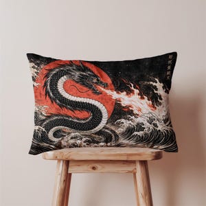 May include: Decorative pillow featuring a black dragon with a white underbelly, set against a red sun and stormy waves. The dragon breathes fire, with Japanese text in the upper right corner. The pillow is rectangular and rests on a wooden stool.