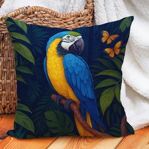 May include: Square decorative pillow featuring a vibrant illustration of a blue and yellow macaw perched on a branch. The pillow has a dark blue background with green leaves and two orange butterflies. The pillow is placed on a wooden surface.