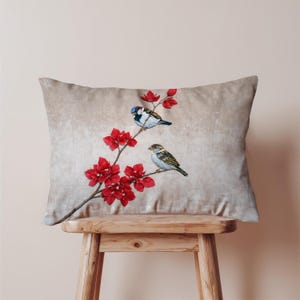 May include: A rectangular cushion with a detailed illustration of two birds perched on a branch with vibrant red flowers. The birds have blue, white, and brown feathers. The cushion has a neutral beige background.