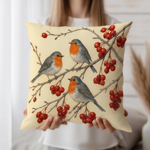 May include: Square decorative pillow featuring a watercolor illustration of three robins perched on branches with red berries. The birds have orange breasts and gray wings against a cream-colored background. The pillow is held in front of a person.