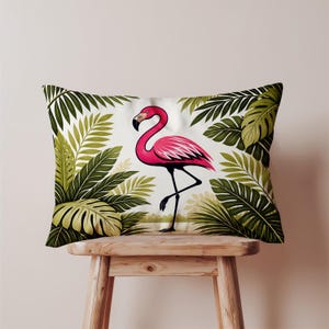 Puede incluir: Cojín rectangular con una ilustración de un flamenco rosa. El flamenco está de pie sobre una pata, rodeado de hojas tropicales verdes. El cojín está colocado sobre un taburete de madera, creando un tema tropical.