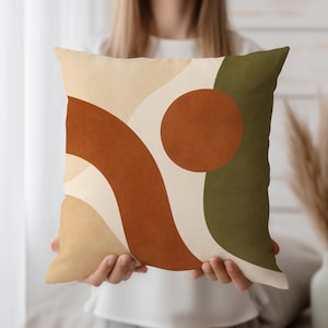 Könnte beinhalten: Quadratisches Zierkissen mit abstraktem Design. Das Kissen zeigt ein Muster aus geschwungenen Linien und einem Kreis in Braun-, Beige- und Olivgrüntönen. Das Kissen wird von einer Person gehalten.