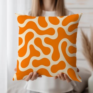 May include: Square decorative pillow with an abstract pattern. The pillow features a cream-colored background with a repeating pattern of orange, amoeba-like shapes. The pillow is a square shape.